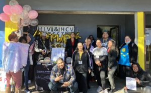 A group of people standing in front of a building, posing and smiling, with balloons and one person wearing a winged PrEP fairy costume